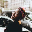 A joyful woman wearing a colorful headwrap and distinctive earrings, smiling in an urban setting.